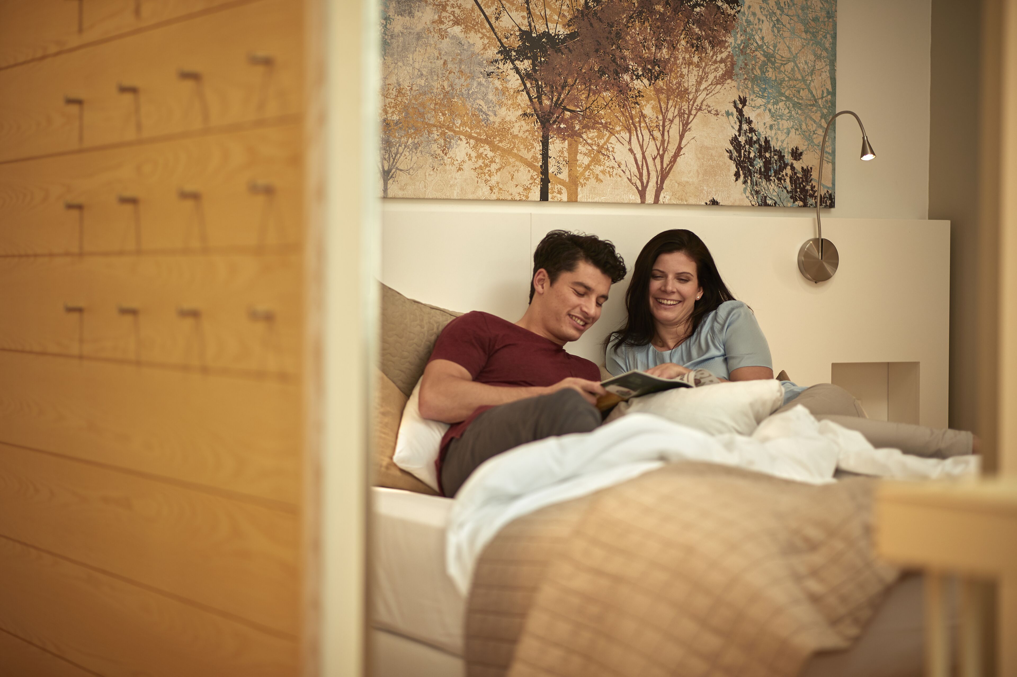 Una pareja sentada en una cama en una habitación con un cuadro grande de árboles en la pared, una lámpara de noche y un armario de madera.