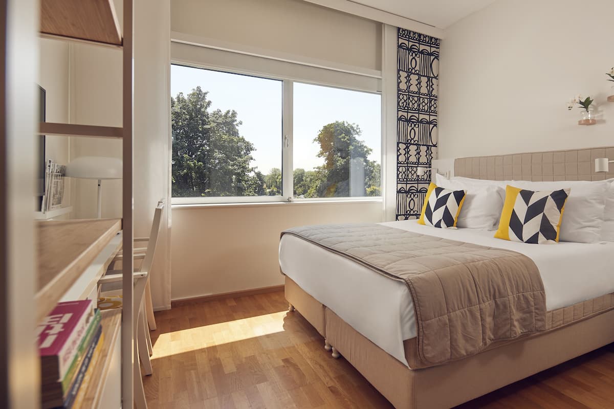 A bedroom with a large bed, a window with a view of trees, a bookshelf, and a bedside table with a lamp.