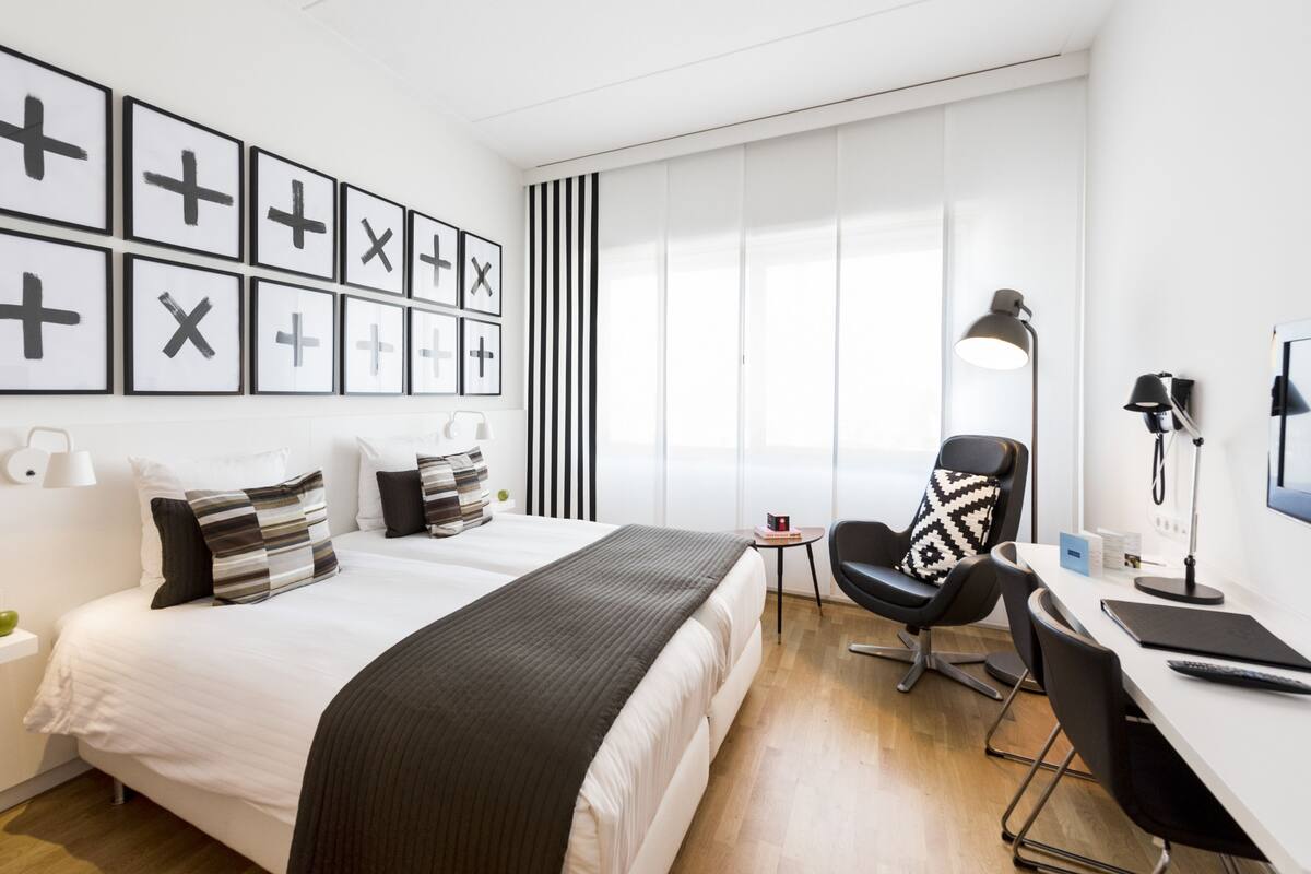 A modern hotel room with a large bed, a desk with a chair, a standing lamp, and a window with blinds.