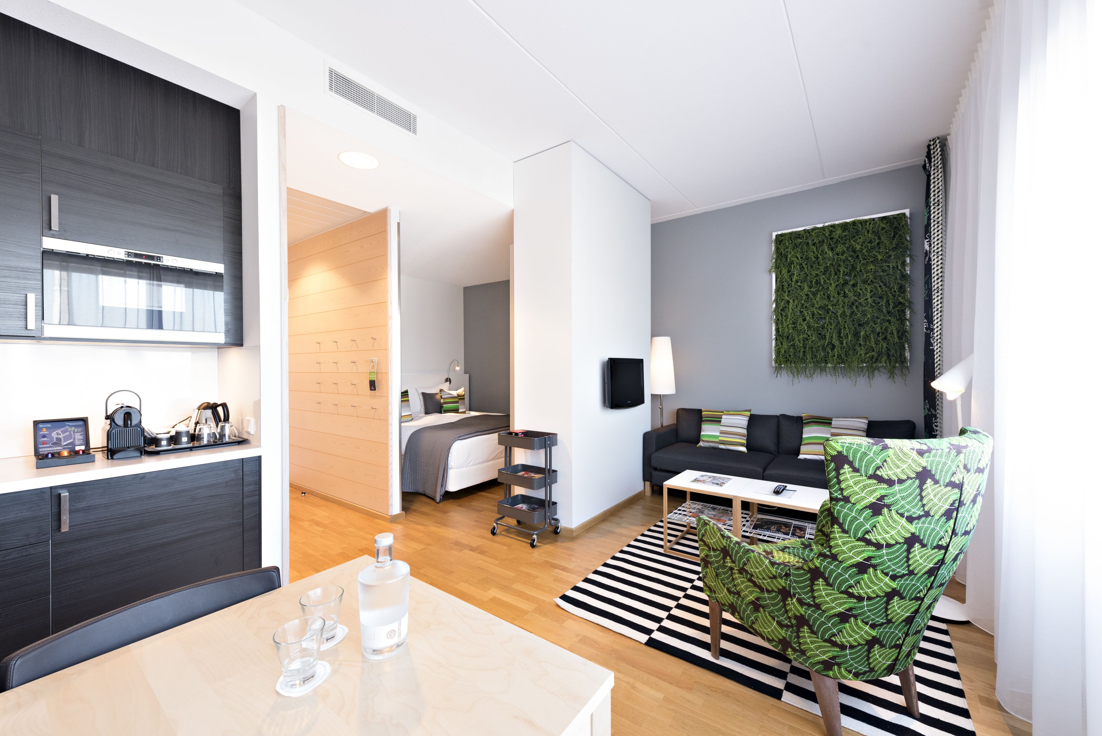 Une chambre d’hôtel moderne dotée d’une kitchenette, d’un coin repas, d’un espace de vie avec un canapé et un fauteuil à motifs verts, ainsi que d’une grande fenêtre agrémentée d’une décoration murale verte.