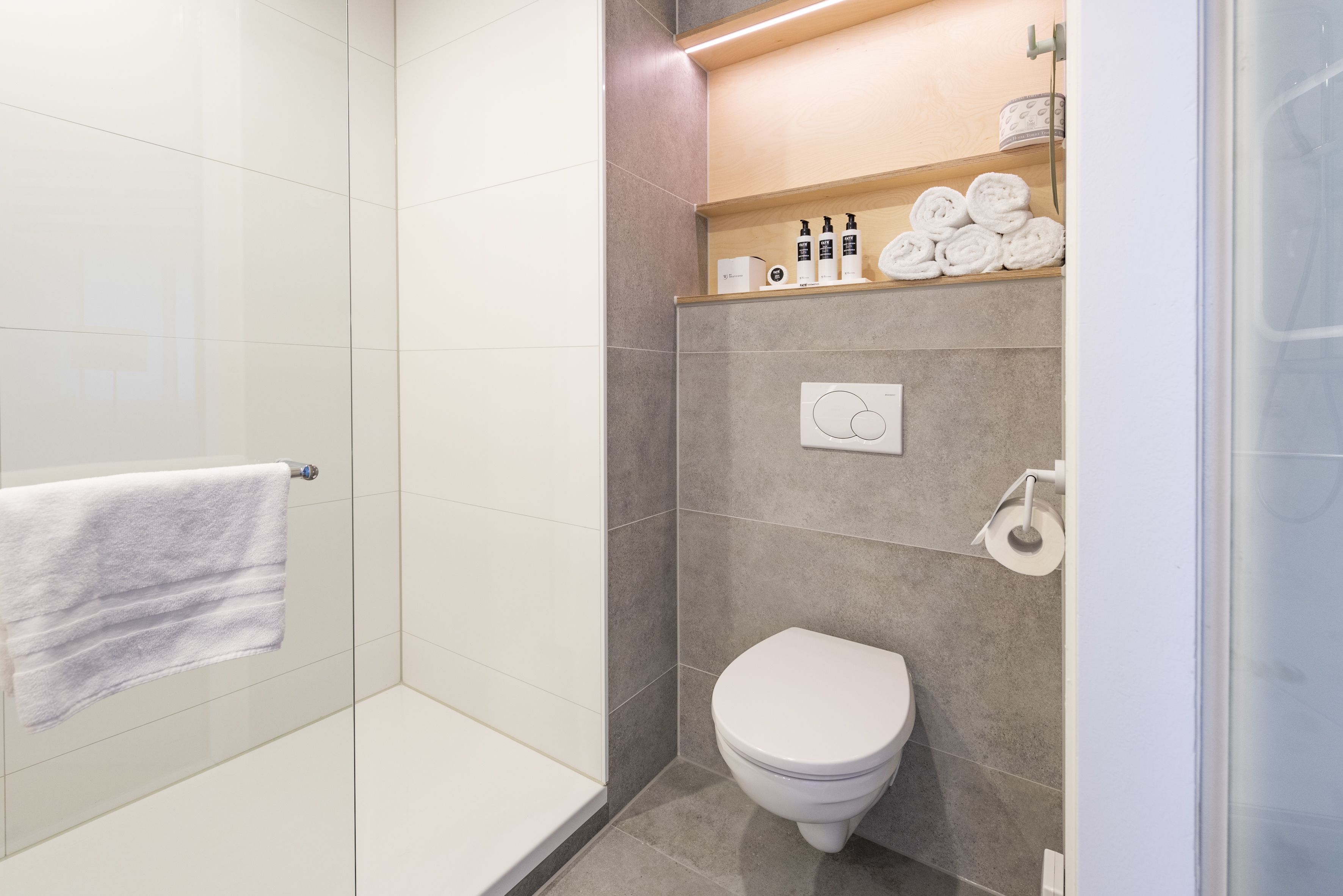 A modern bathroom with a glass shower enclosure, a white toilet, and a shelf with toiletries.