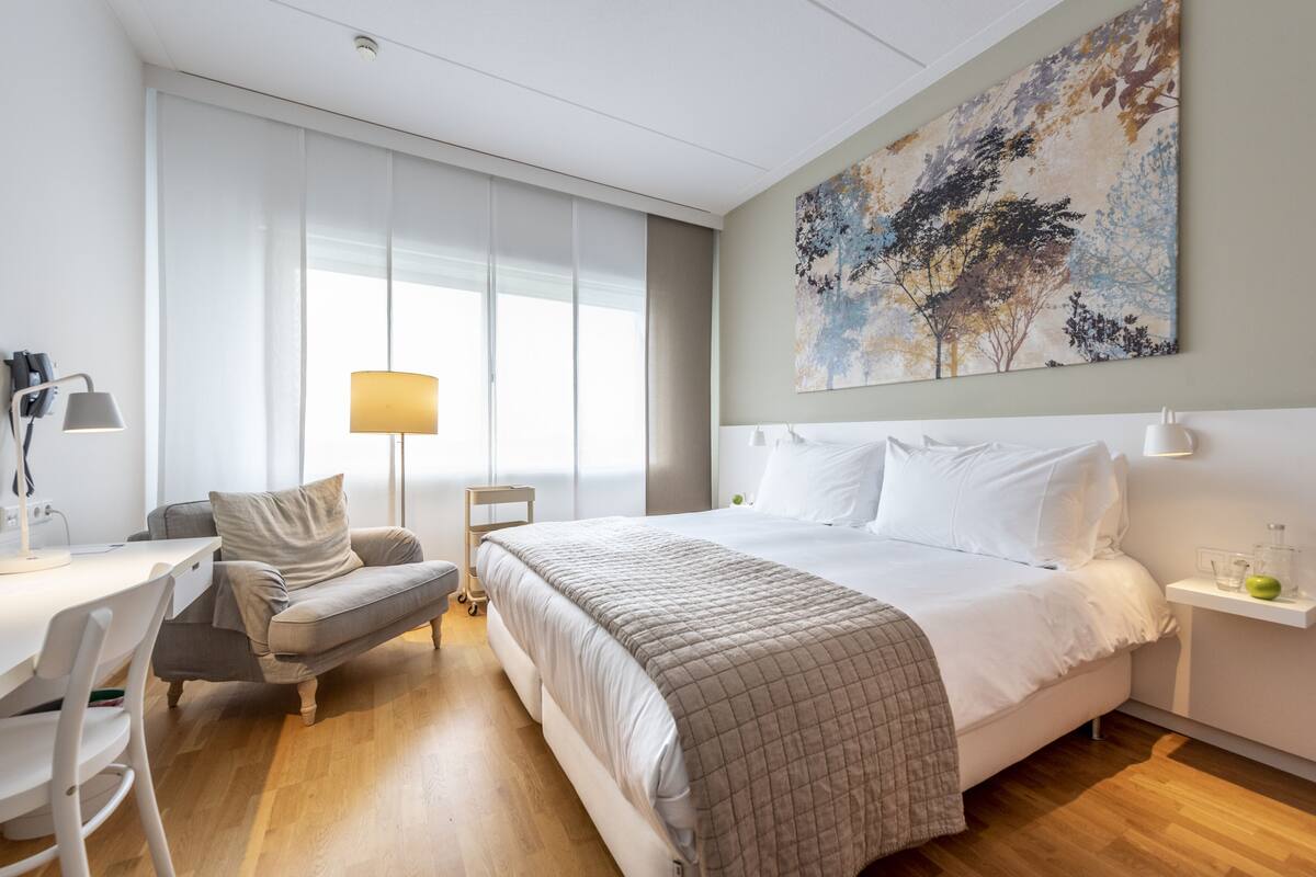A modern hotel room with a large bed, a desk, a chair, and a lamp.