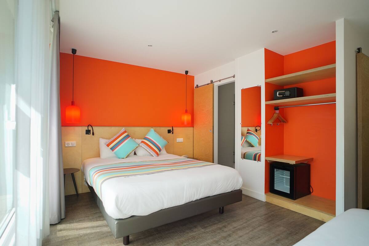 A modern hotel room with a large bed, orange walls, and a built-in wardrobe with shelves.