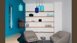 A modern living room with teal walls, a wooden coffee table, two patterned armchairs, a small side table, and a bookshelf with various items.