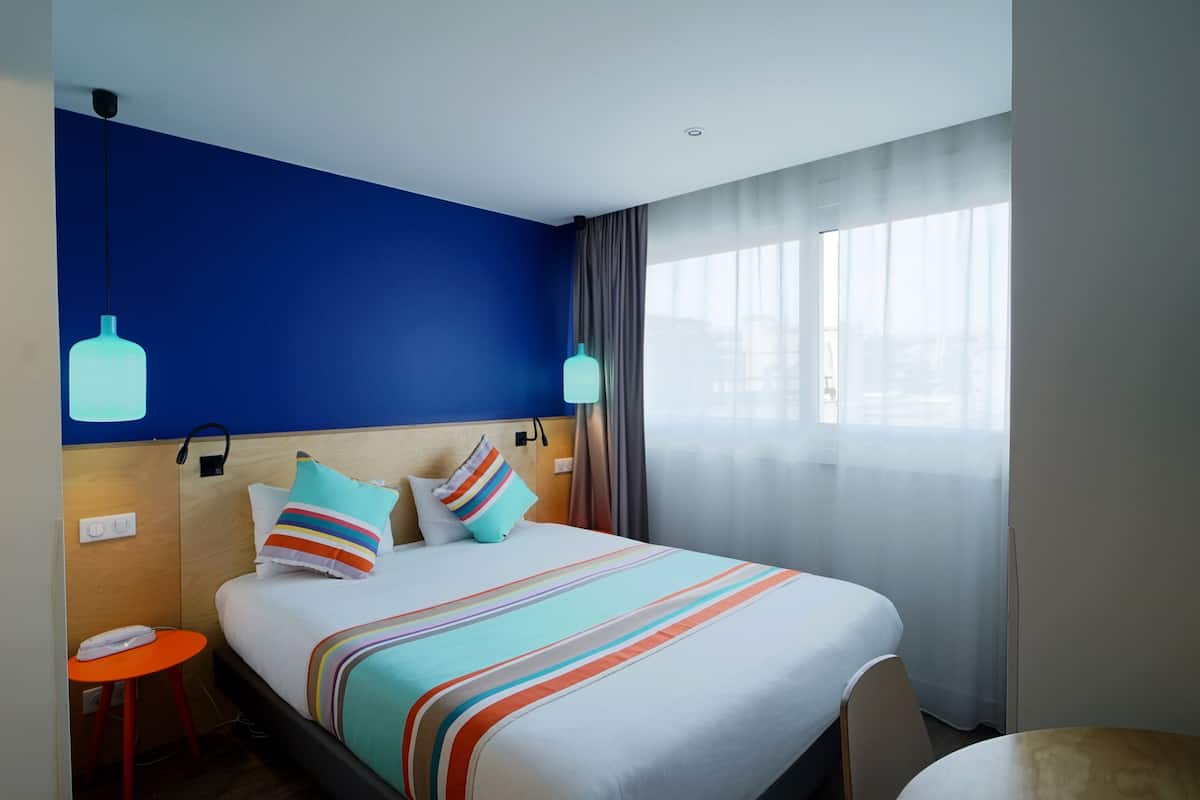 A hotel room with a large bed, two bedside tables, a nightstand, a window with sheer curtains, and a blue accent wall.