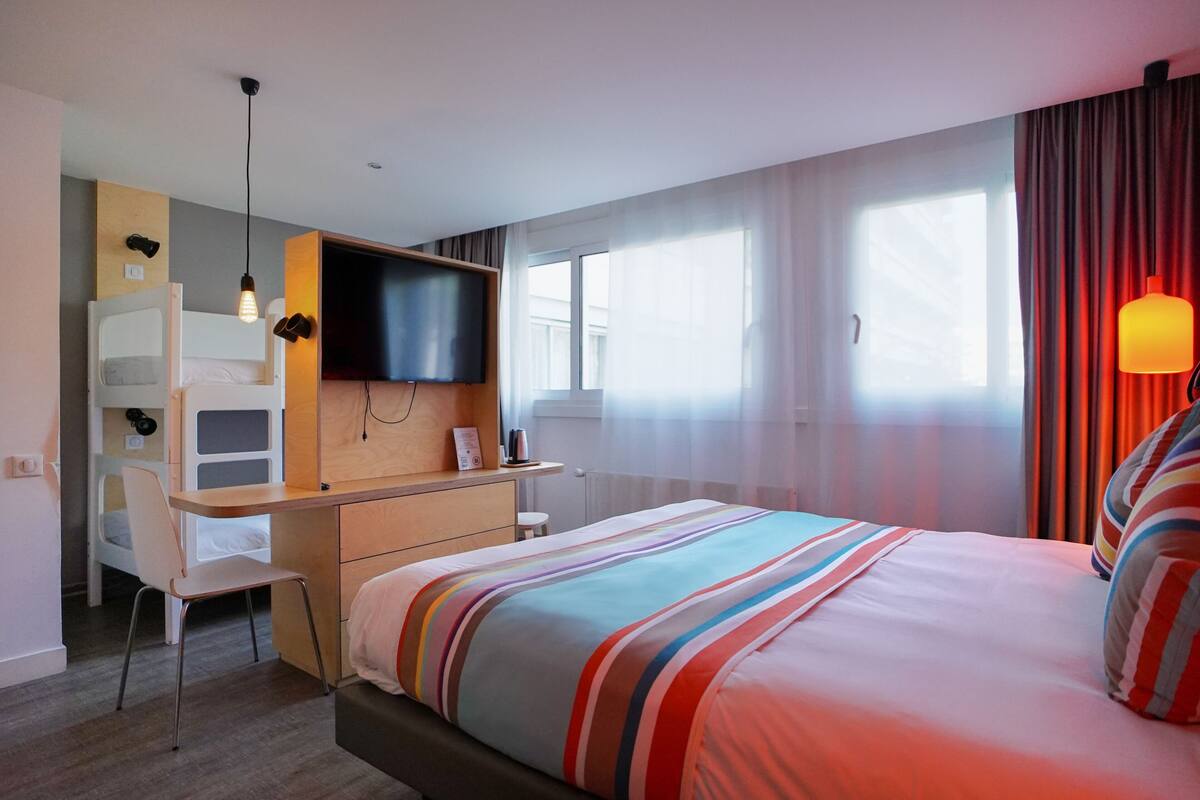 A hotel room with a bed, a desk with a chair, a television, and a window with curtains.