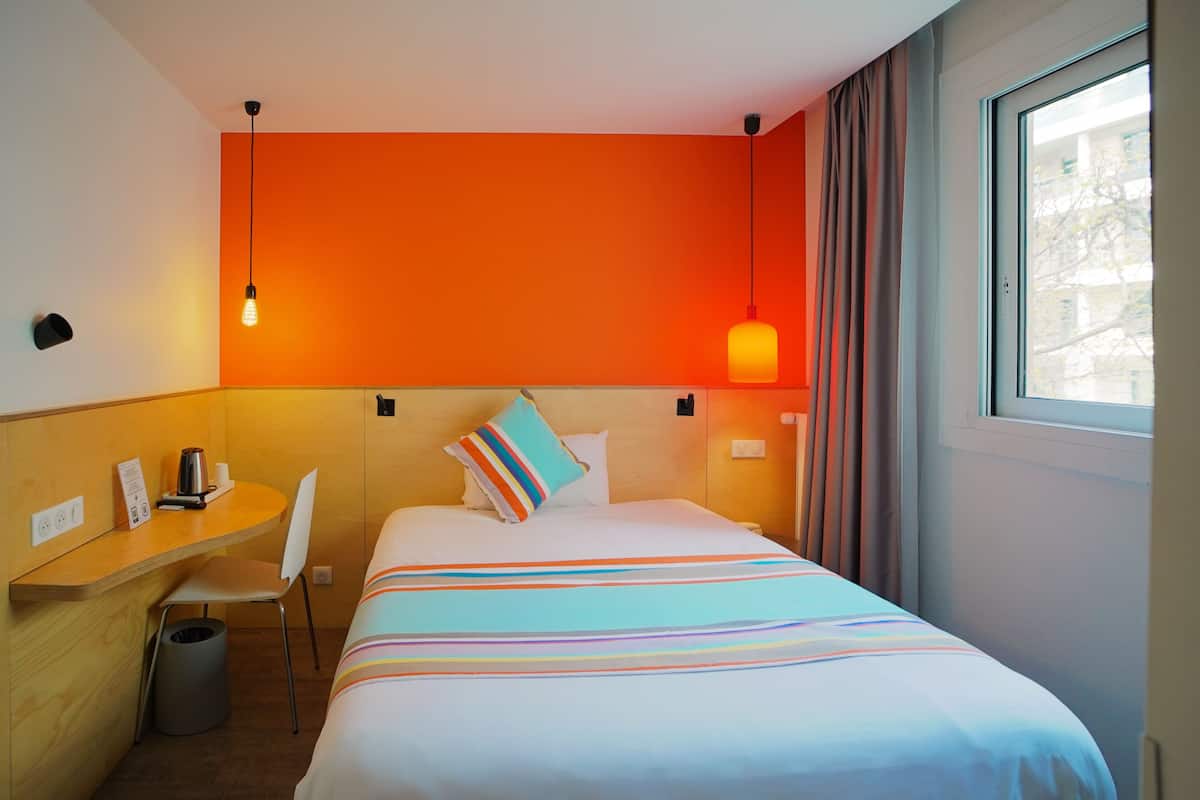 A hotel room with a bed, a desk, a chair, a trash bin, a window, and a wall with a light fixture.