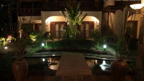 Um deck de madeira leva a um lago com uma pequena ponte, cercado por vegetação exuberante e um edifício com varanda ao fundo.