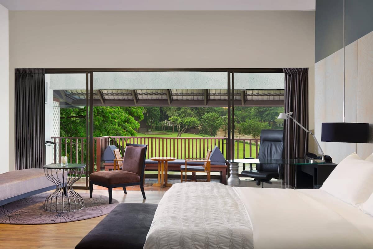 Une chambre d’hôtel moderne dotée d’un grand lit, d’un bureau, d’une chaise et offrant une vue sur un jardin luxuriant grâce à de grandes fenêtres.