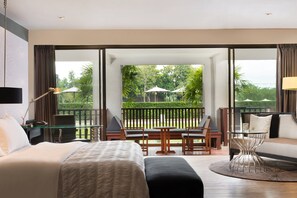 A modern hotel room with a large bed, a desk, and a sofa. There is a balcony with outdoor furniture and a view of greenery.
