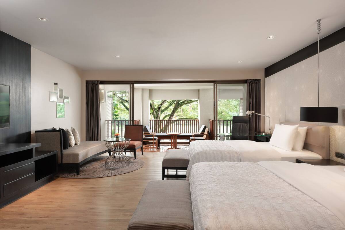 Une chambre d’hôtel moderne avec deux lits, un coin salon avec une table et des chaises, une télévision et un balcon offrant une vue sur les arbres.