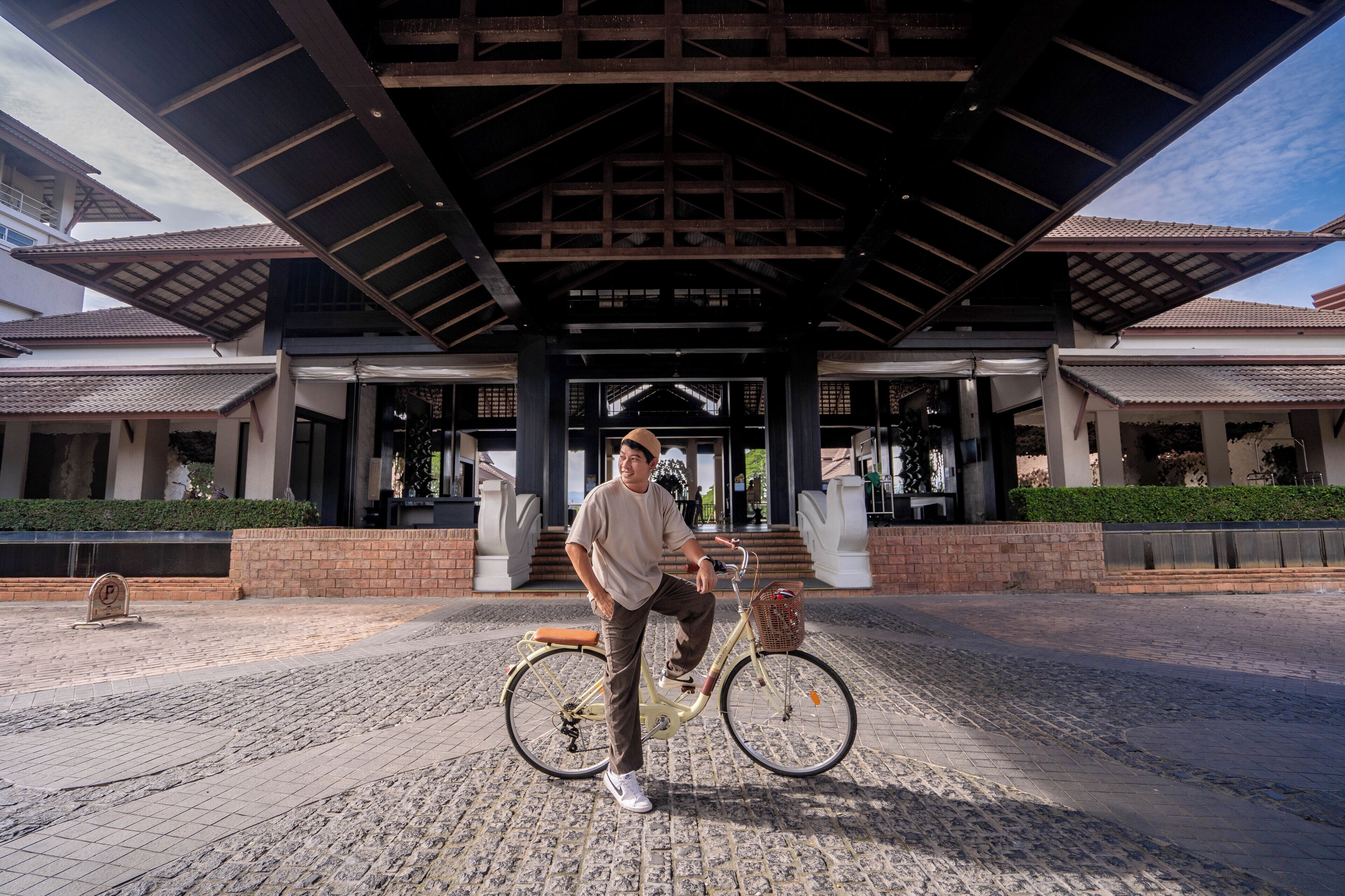 A person standing next to a bicycle in front of a building with a covered entrance and a tiled roof.
