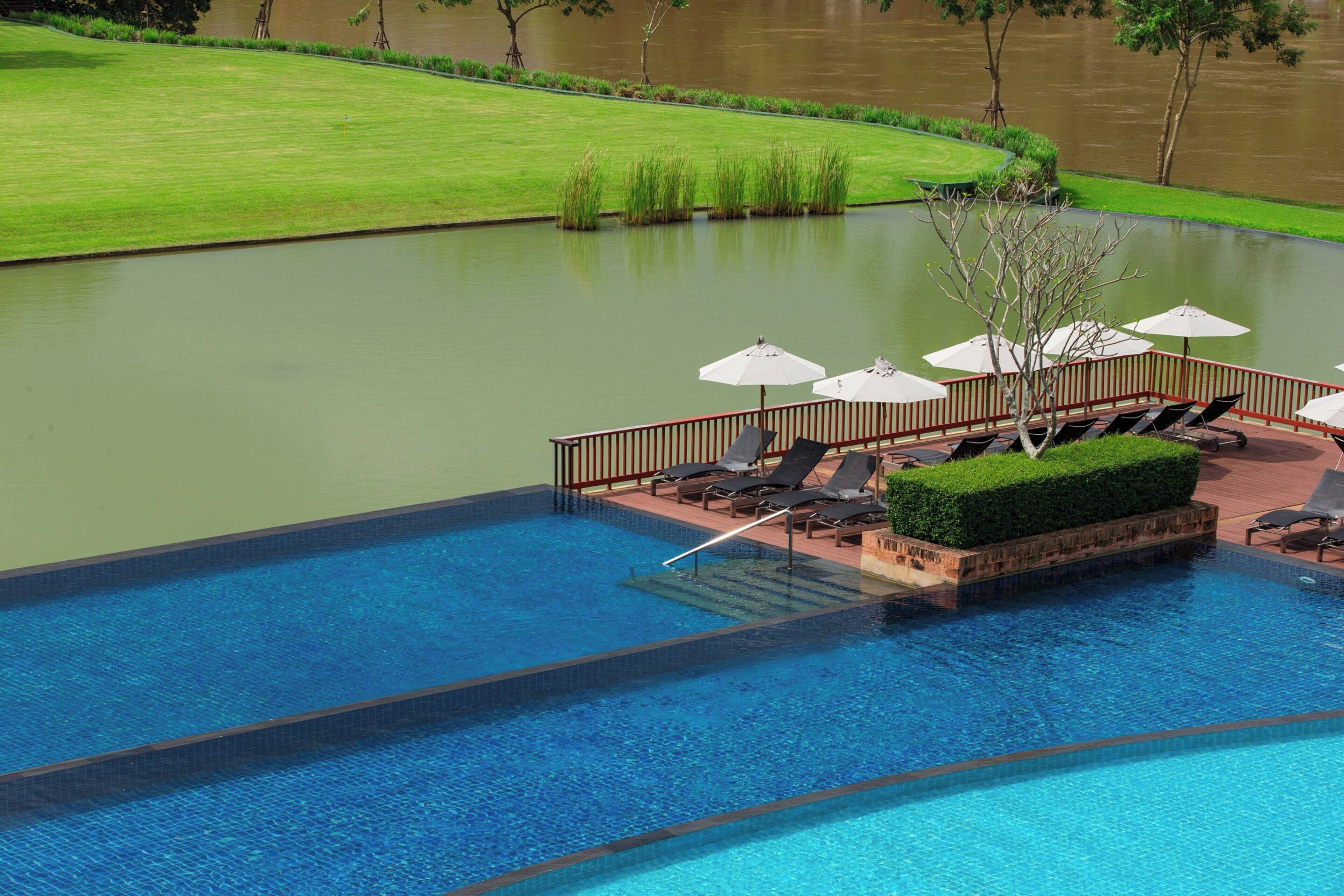A swimming pool with lounge chairs and umbrellas, surrounded by a landscaped area and a view of a river.