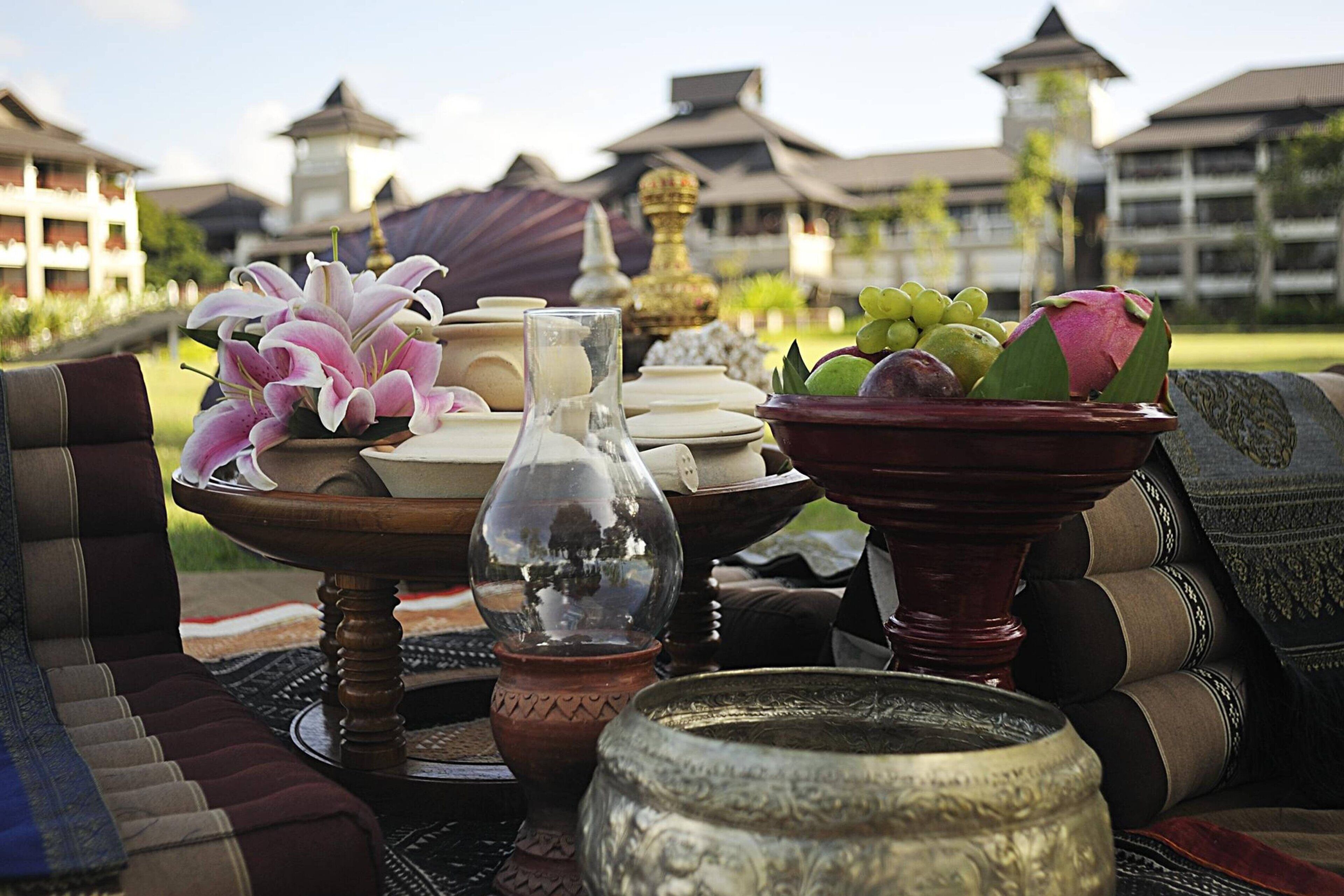 A table set with traditional Asian-style tableware, including a vase of pink lilies, a glass pitcher, and a bowl of fruit, with a view of traditional buildings in the background.