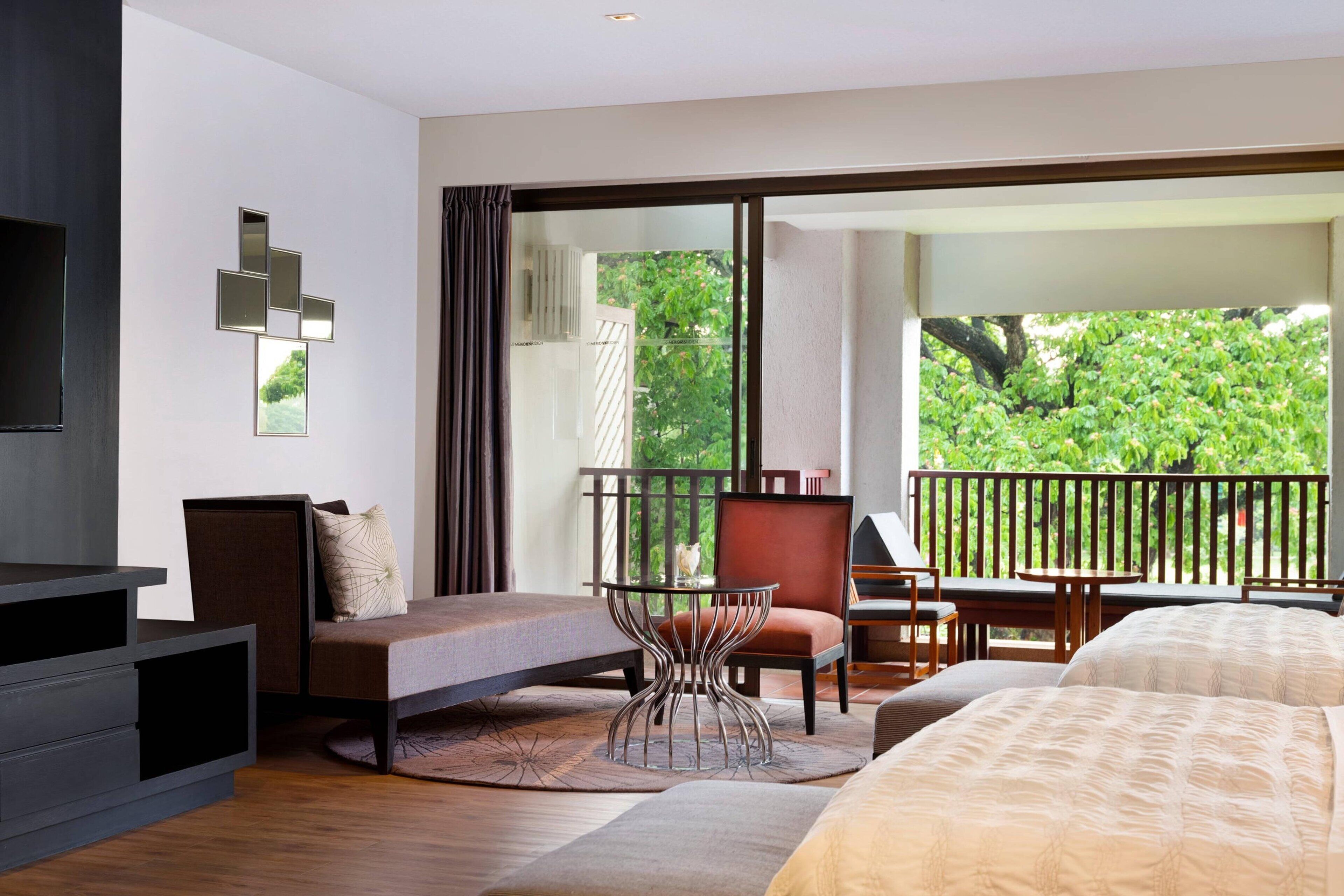 Una habitación de hotel moderna con una cama grande, un área de descanso con una mesa y un balcón con vistas a zonas verdes.