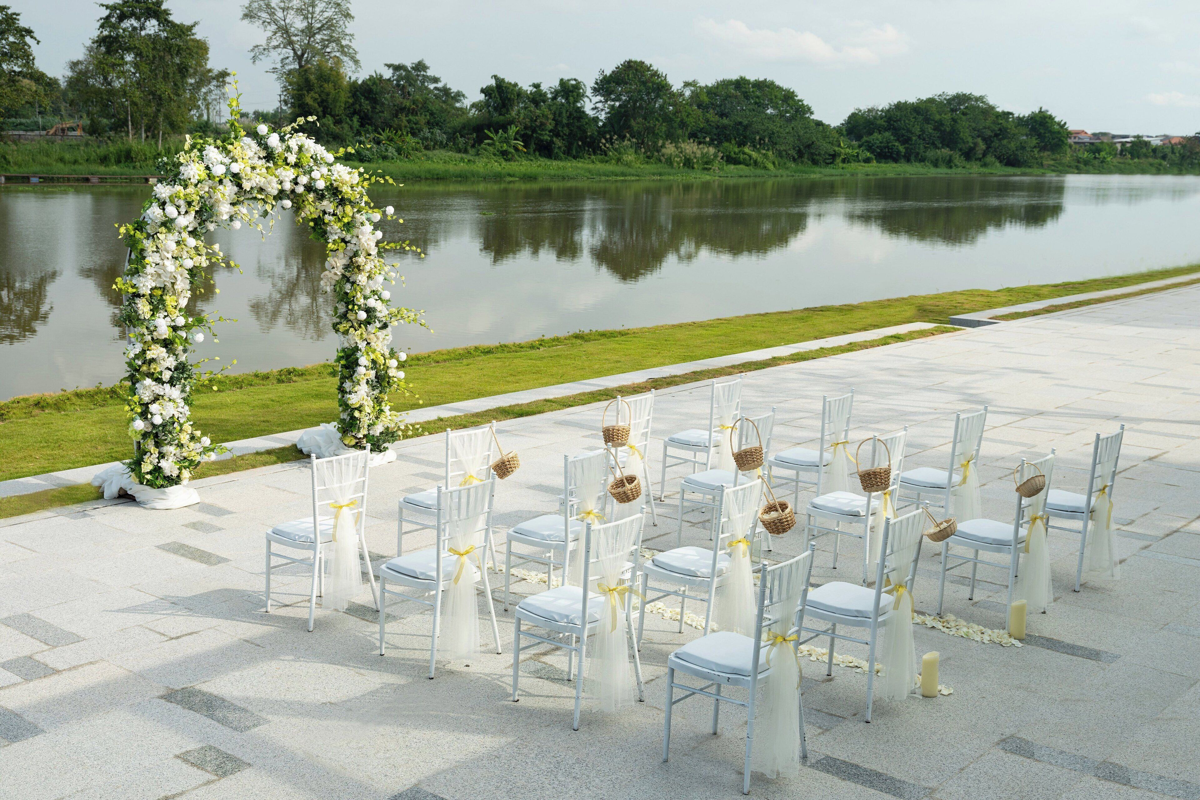 A wedding ceremony setup with chairs arranged in rows, a floral arch, and a calm lake in the background.
