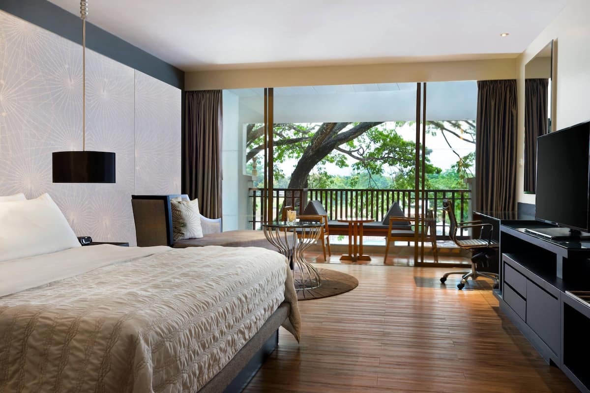 Une chambre d’hôtel moderne dotée d’un grand lit, d’une télévision, d’un coin repas avec une table et des chaises, et d’un balcon offrant une vue sur les arbres.