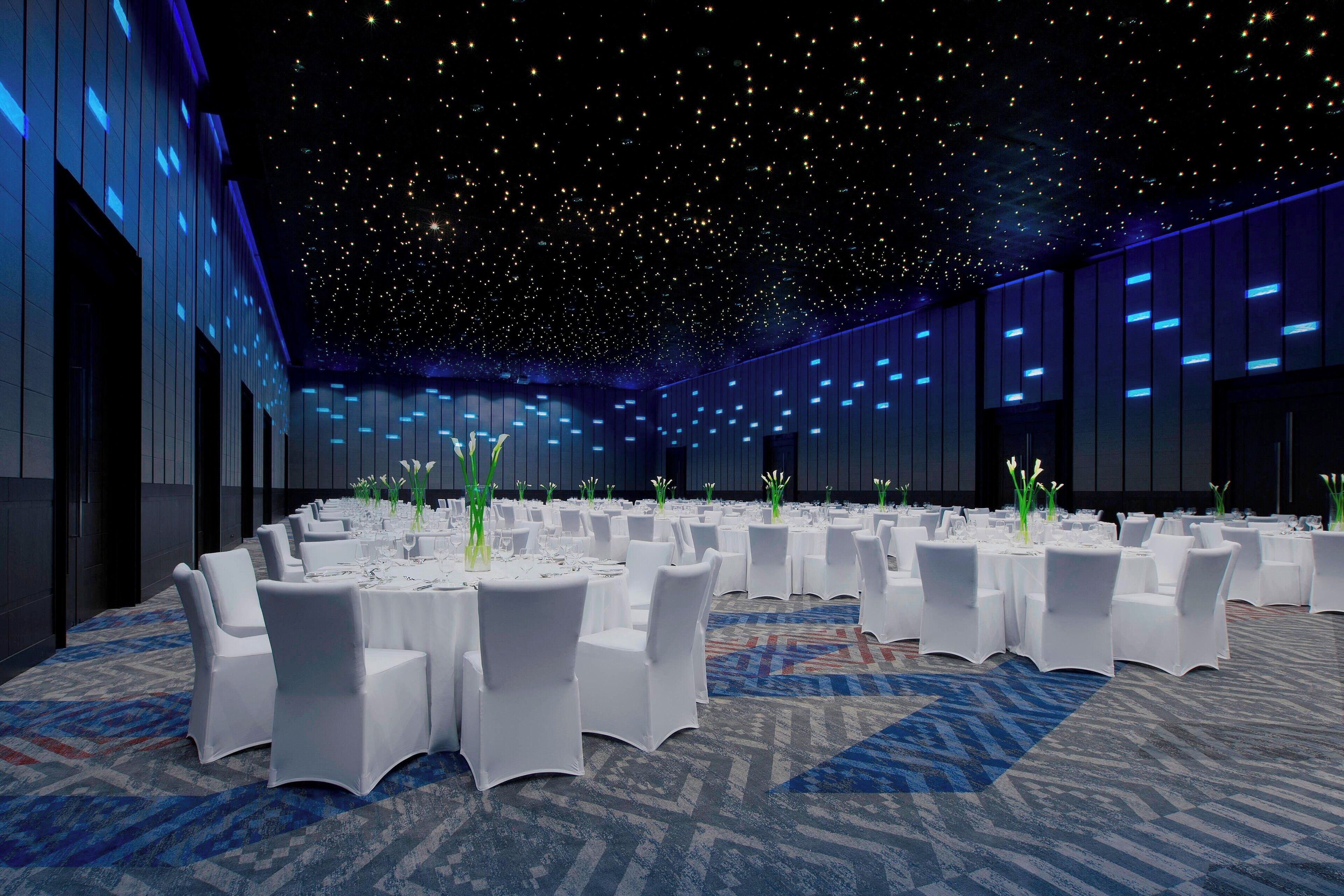 A large conference room with round tables and white chairs, decorated with flowers and set for a formal event.