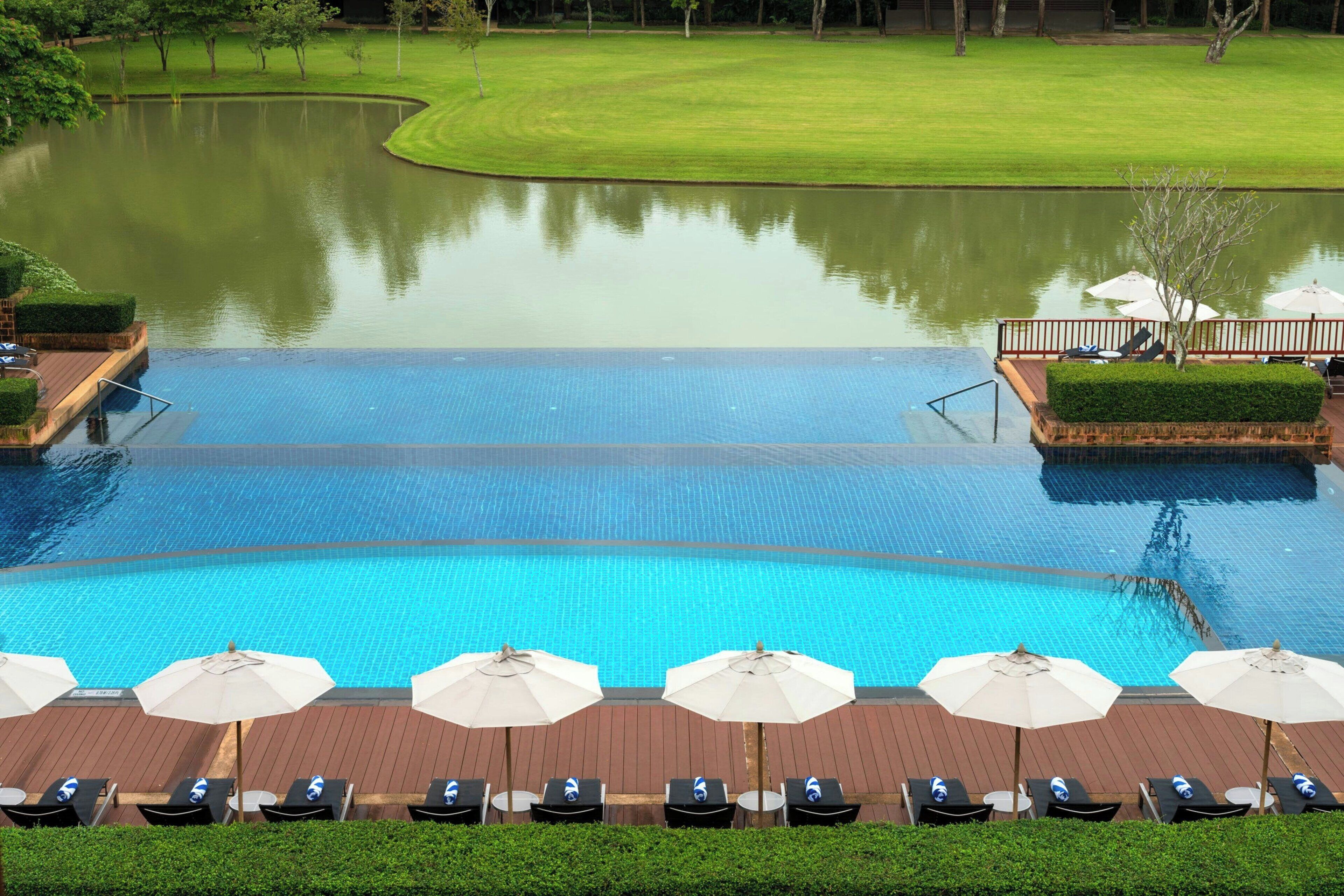 Una gran piscina con sillones de playa y sombrillas, rodeada de un área ajardinada con un estanque y un campo de golf al fondo.