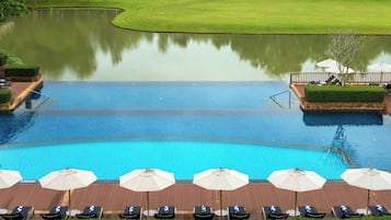 Uma grande piscina com espreguiçadeiras e guarda-sóis, rodeada por um jardim paisagístico com lago e um campo de golfe ao fundo.
