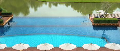 Una gran piscina con sillones de playa y sombrillas, rodeada de un área ajardinada con un estanque y un campo de golf al fondo.