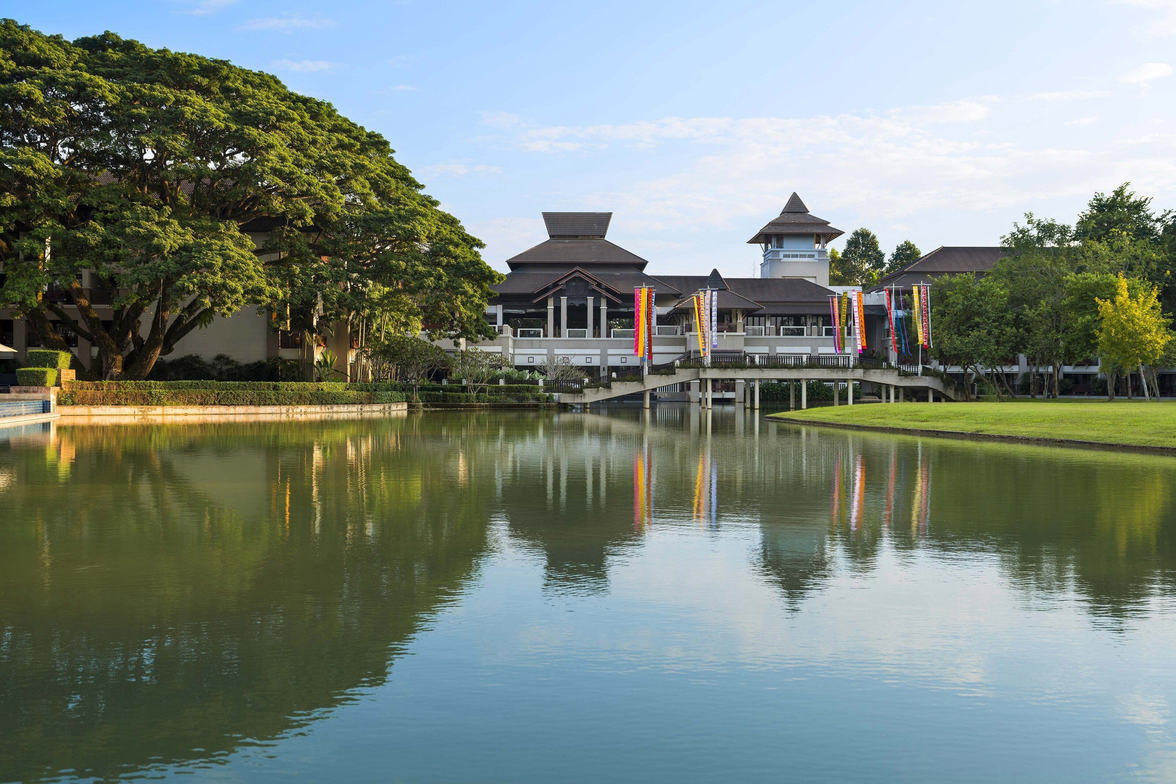 A traditional building with a reflection in a calm water body, surrounded by greenery and a clear sky.