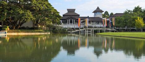 A traditional building with a reflection in a calm water body, surrounded by greenery and a clear sky.
