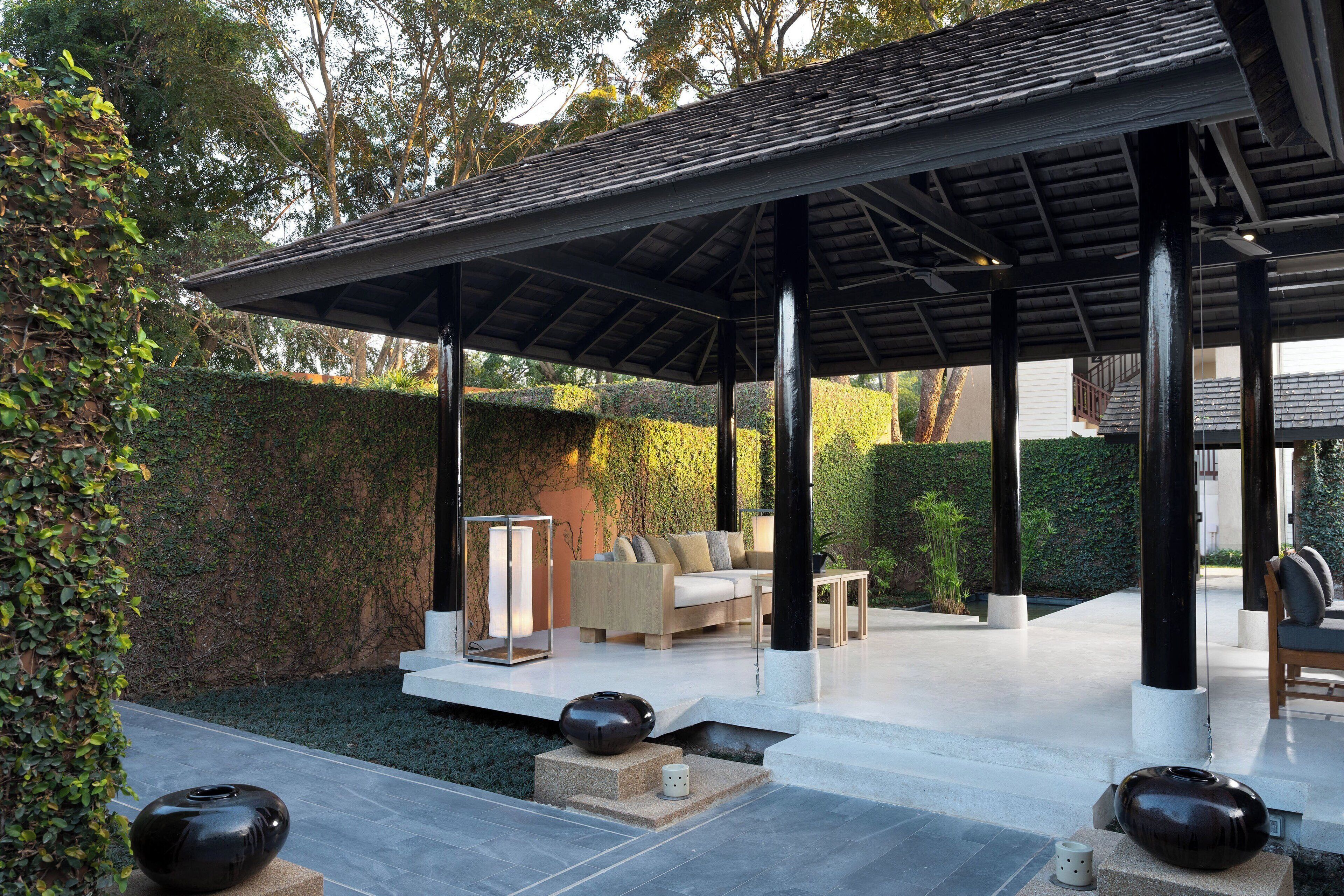A covered outdoor seating area with a sofa and coffee table, surrounded by a hedge and a stone pathway.