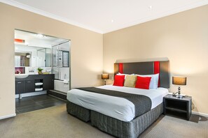 A modern hotel room with a large bed, a bedside table with a lamp, a built-in wardrobe, and a bathroom visible through a glass door. - Platinum International (Harristown)