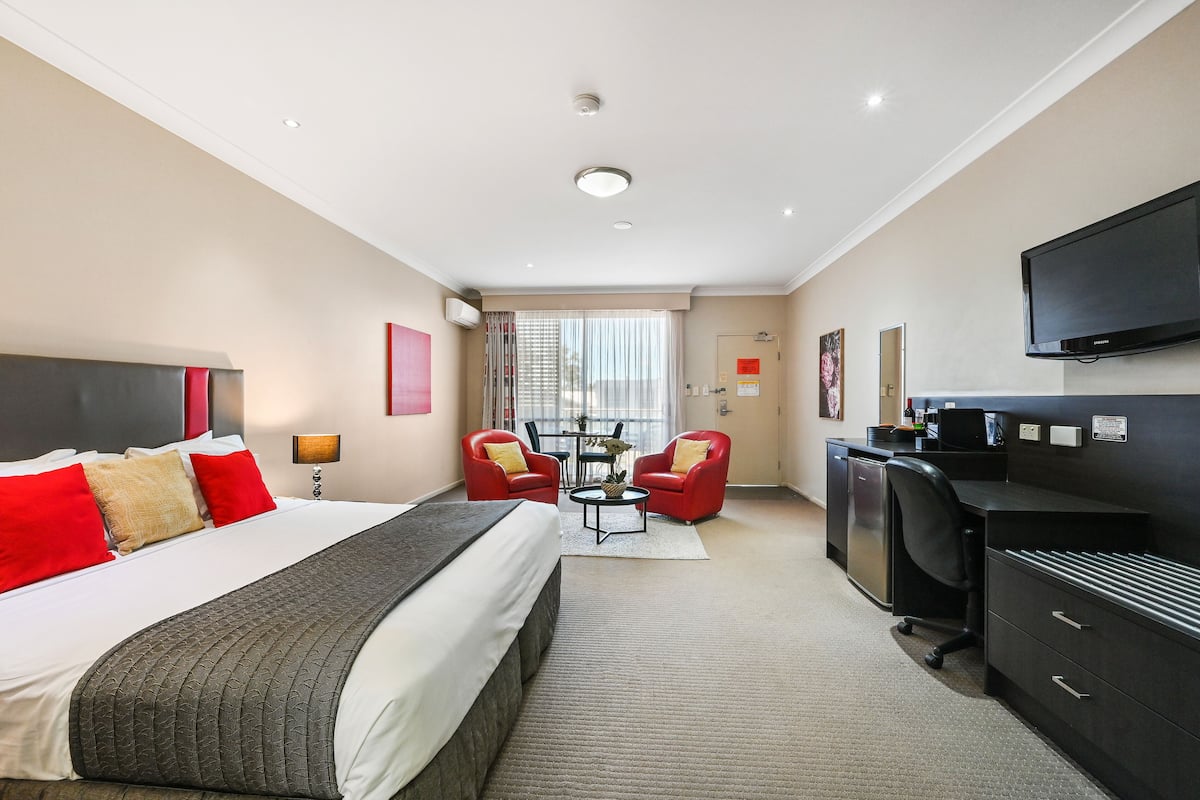 Habitación de hotel con una cama grande, dos sillas rojas, un escritorio, un televisor y una ventana con cortinas.