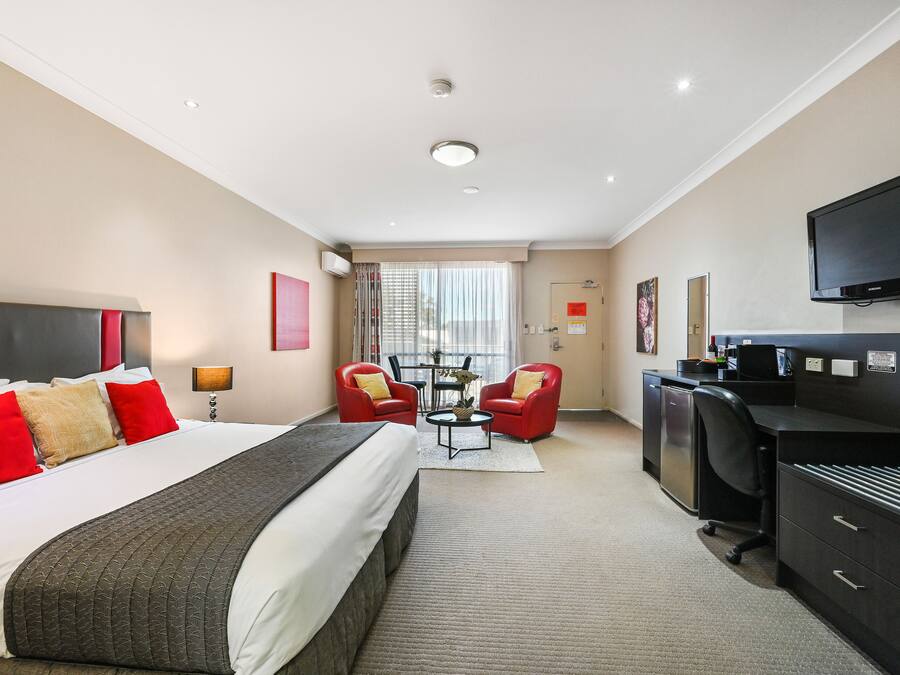 A hotel room with a large bed, two red chairs, a desk, a TV, and a window with curtains.