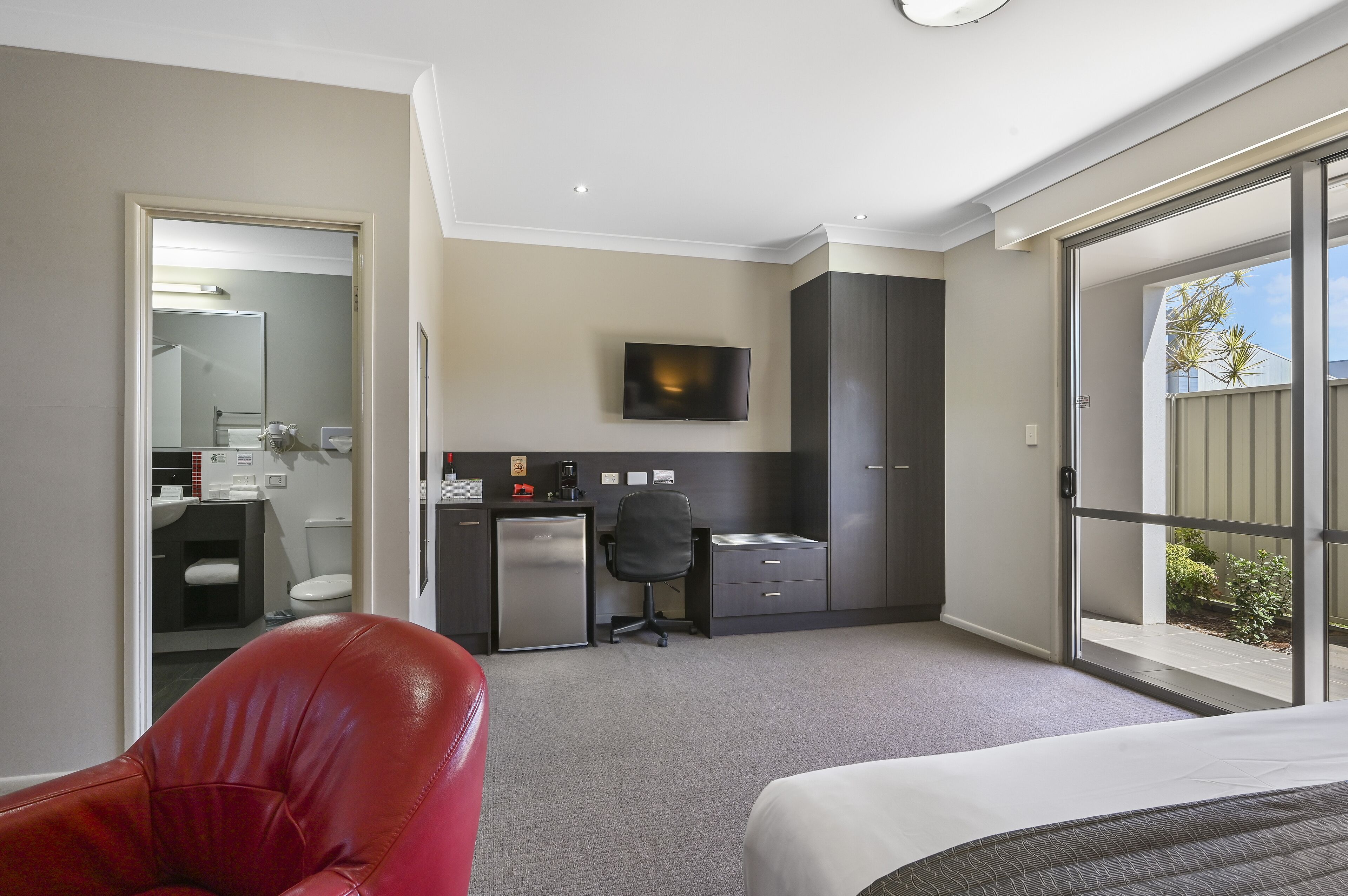 A hotel room with a bed, a red chair, a desk with a computer, a TV, and a balcony with a view.