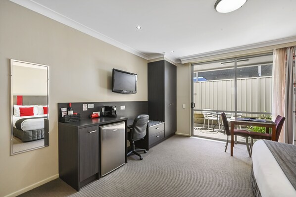 Habitación de hotel con cama, escritorio con computadora, silla, refrigerador pequeño, televisor montado en la pared y balcón con mesa y sillas.
