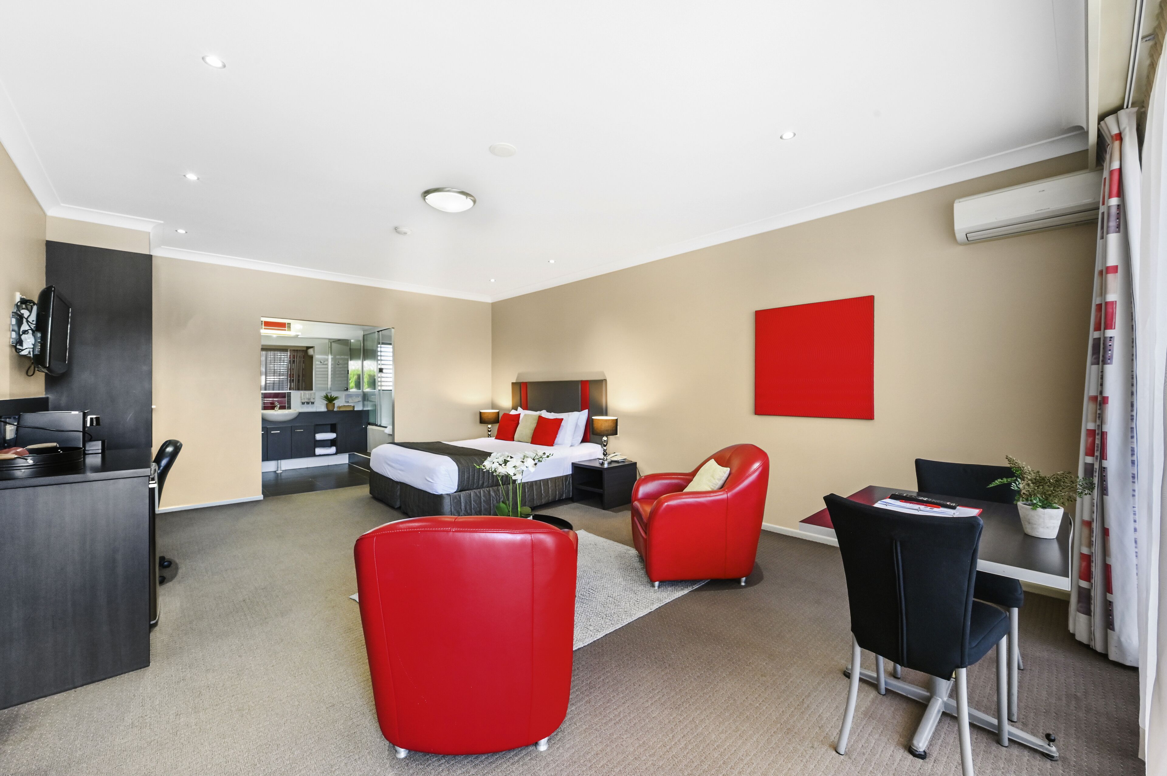 Ein modernes Hotelzimmer mit einem Bett, einem Schreibtisch, einem roten Sessel und einer Essecke mit Tisch und Stühlen.