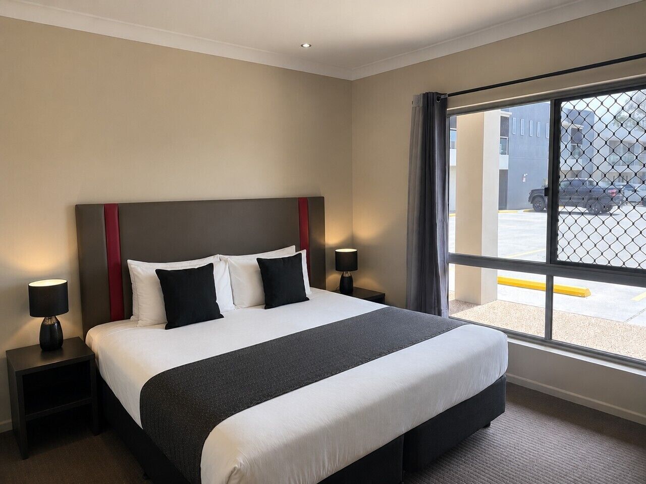 Une chambre d’hôtel avec un grand lit, deux tables de chevet avec des lampes, une fenêtre donnant sur le parking et un détail rouge sur la tête de lit.