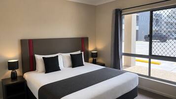 Une chambre d’hôtel avec un grand lit, deux tables de chevet avec des lampes, une fenêtre donnant sur le parking et un détail rouge sur la tête de lit.