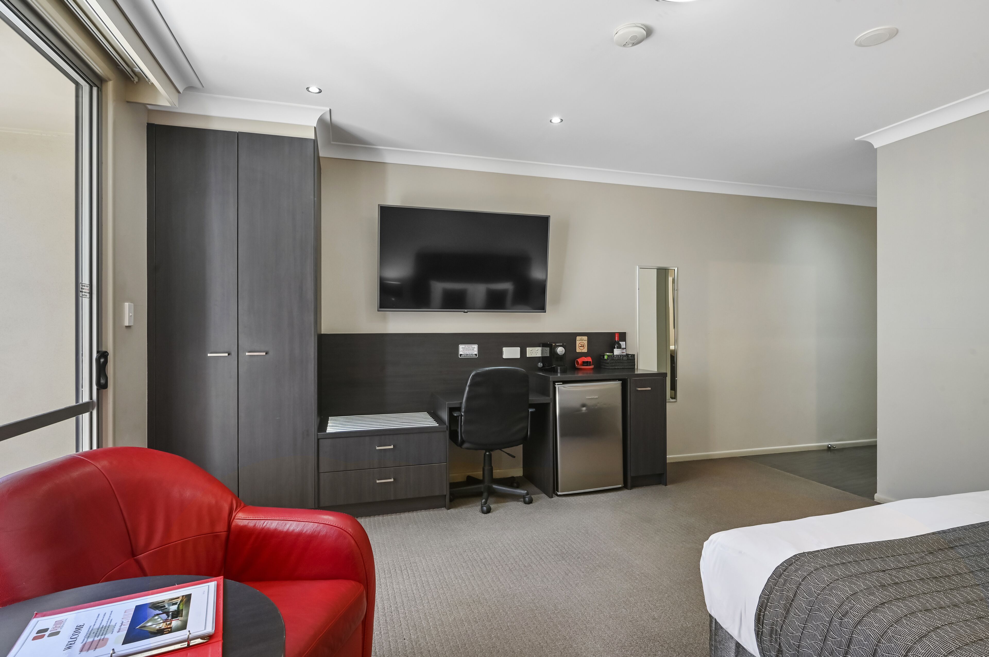Una habitación de hotel con una cama, un escritorio con una silla, un sillón de cuero rojo, un televisor montado en la pared y un minibar.