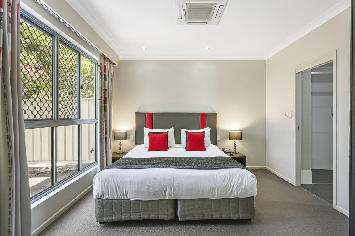 A hotel room with a large bed, two bedside tables with lamps, a window with curtains, and a door to the right.