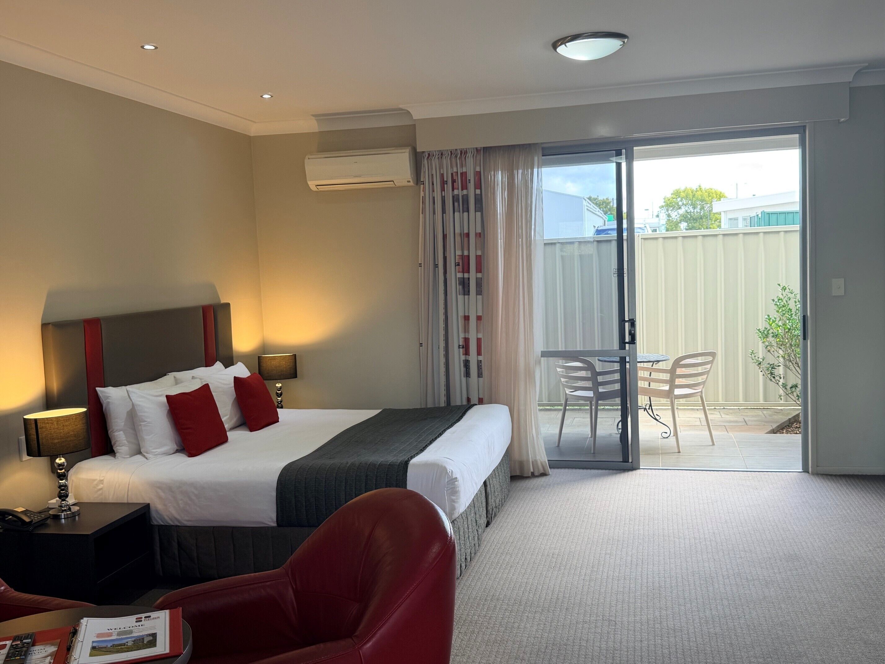 Una habitación de hotel con cama, mesita de noche, lámpara y vistas al balcón.