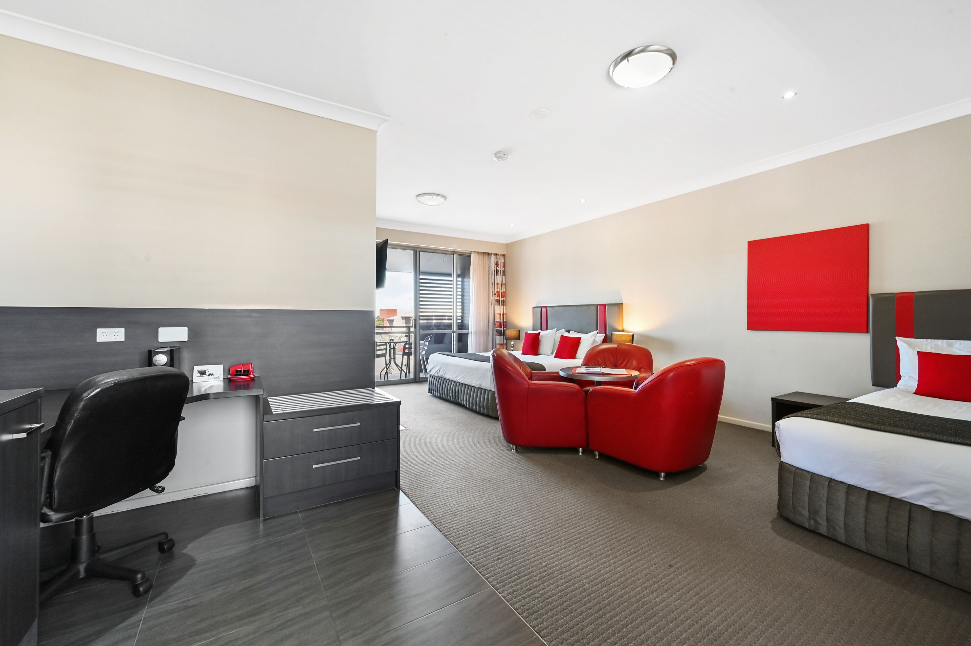 A modern hotel room with a desk, office chair, and a bed with red accents.