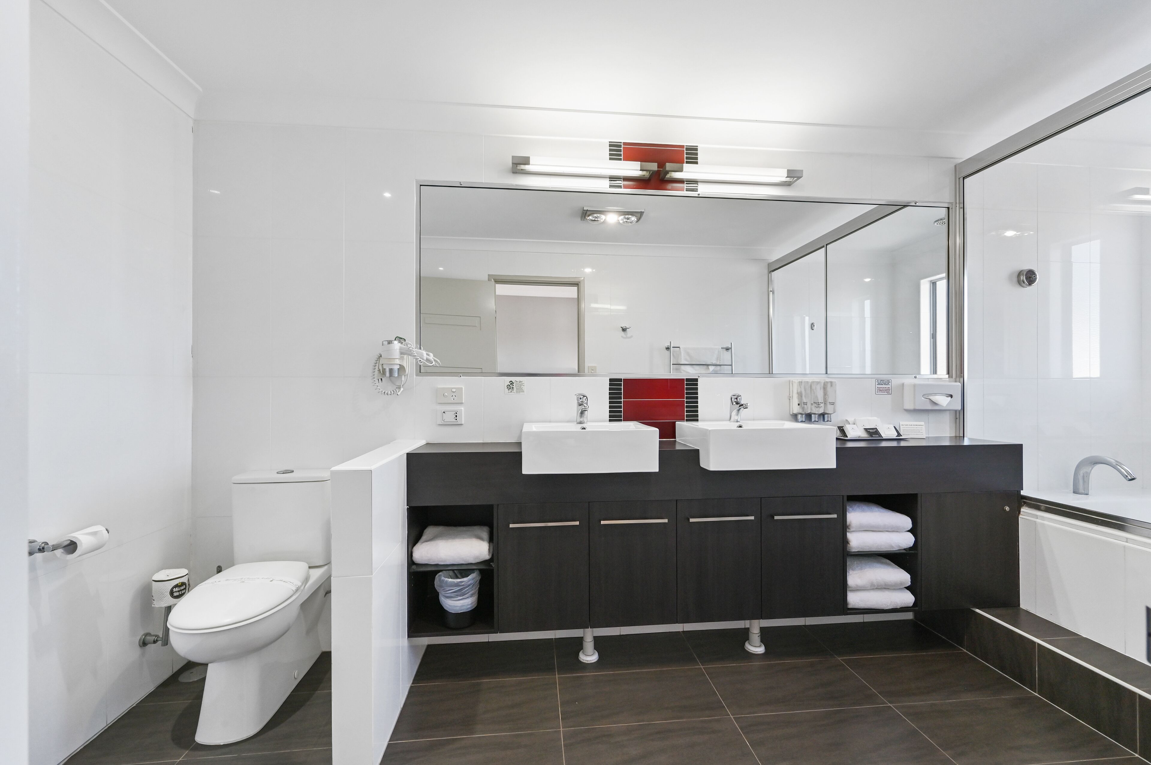 A modern bathroom with a white toilet, dual sinks, a large mirror, and dark cabinetry.