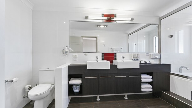 Un baño moderno con inodoro blanco, lavamanos dobles, un espejo grande y muebles oscuros.