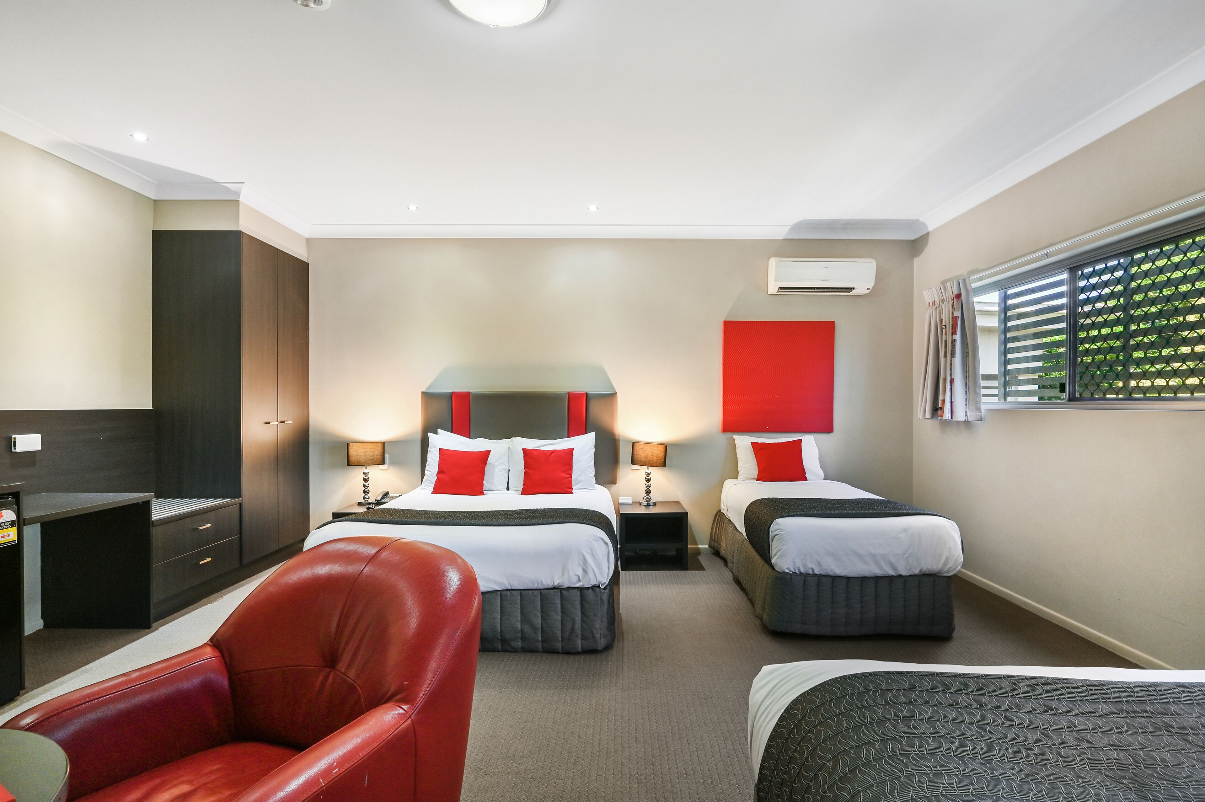 A hotel room with two beds, a red chair, a desk, and a wardrobe.