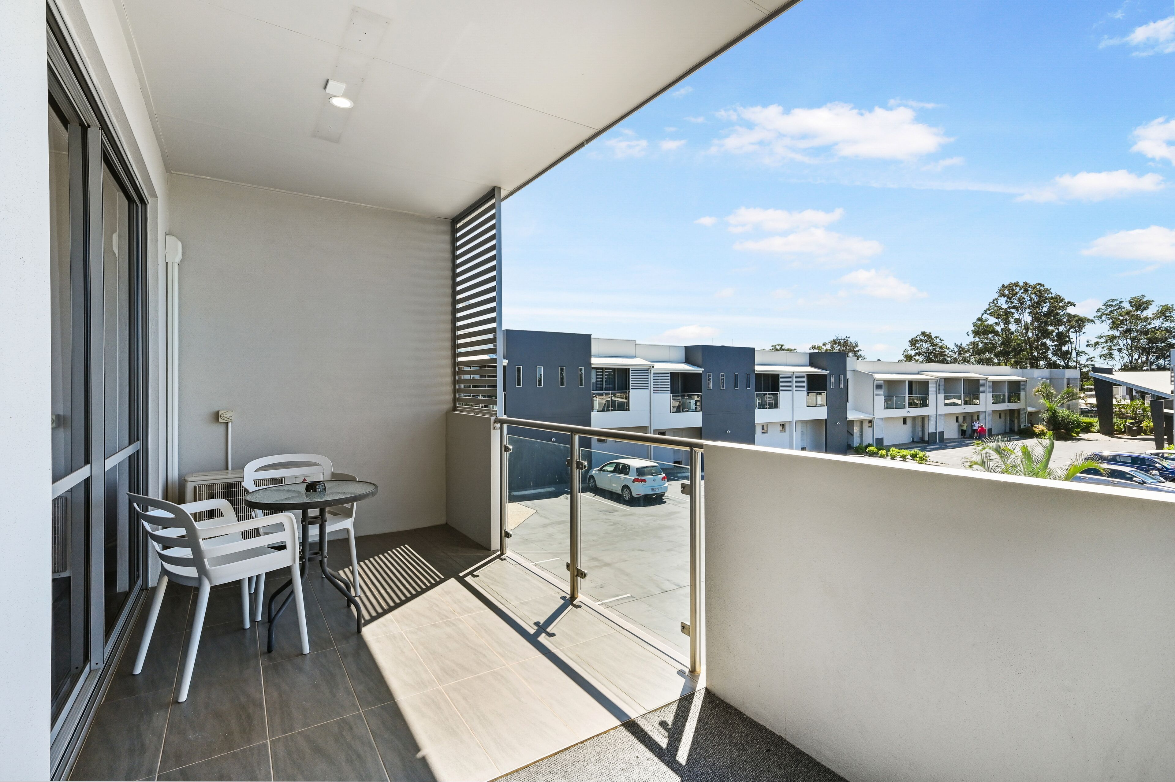 A balcony with a table and chairs, a view of a parking area, and modern buildings in the background.