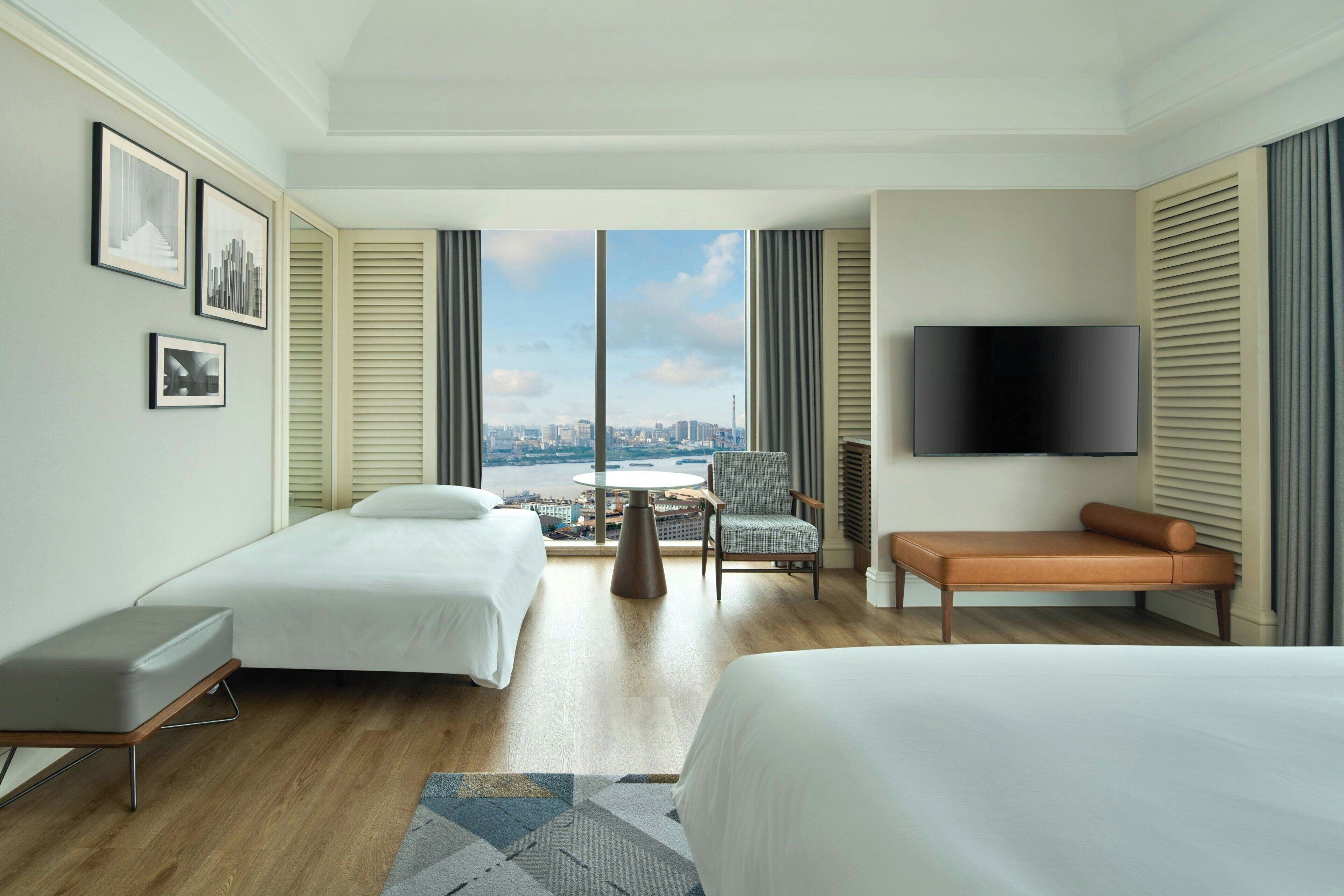 A hotel room with two beds, a TV, a chair, a table, and a view of a cityscape.
