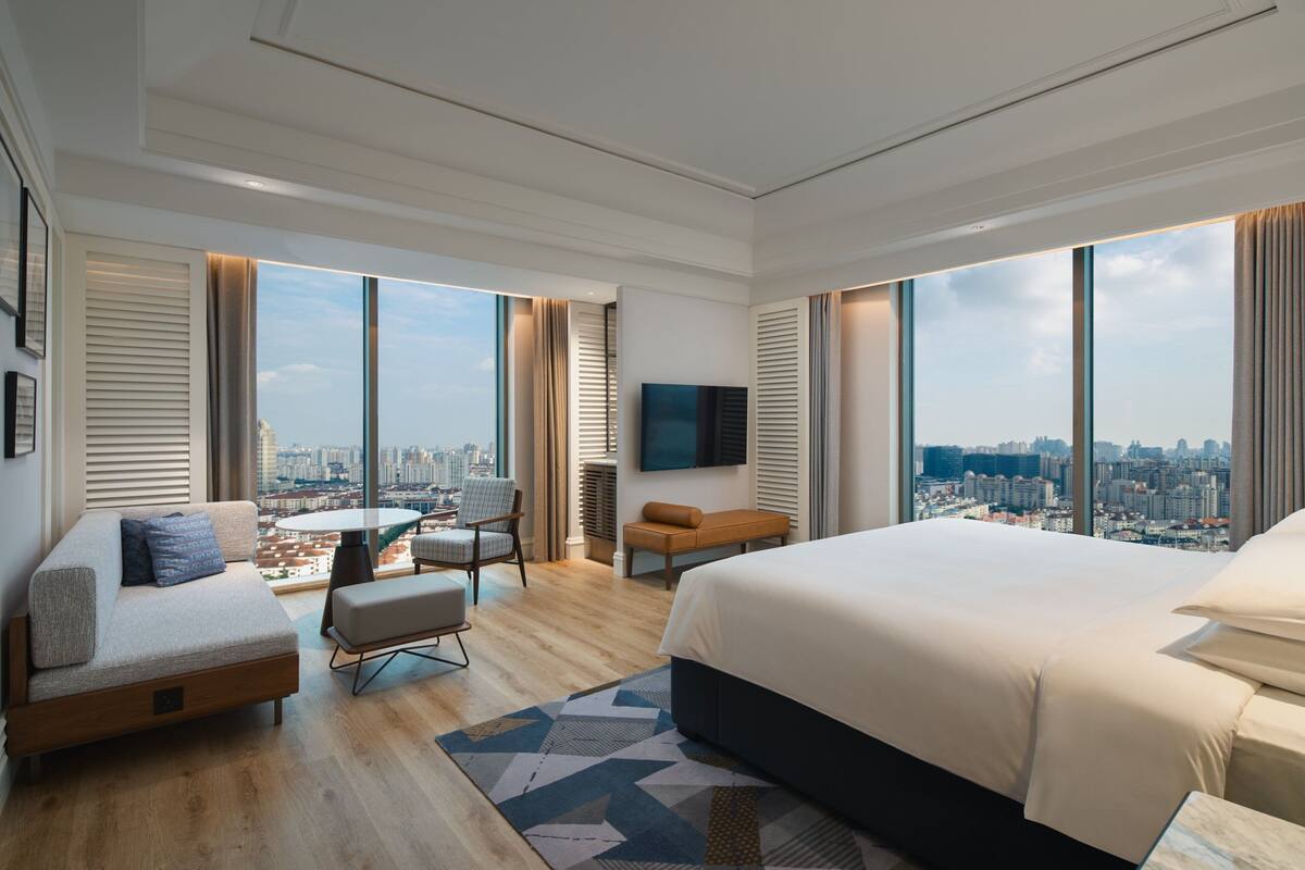 Una habitación de hotel moderna con una cama grande, un sofá, una mesa de comedor y una silla. La habitación tiene ventanas amplias con vistas a la ciudad.