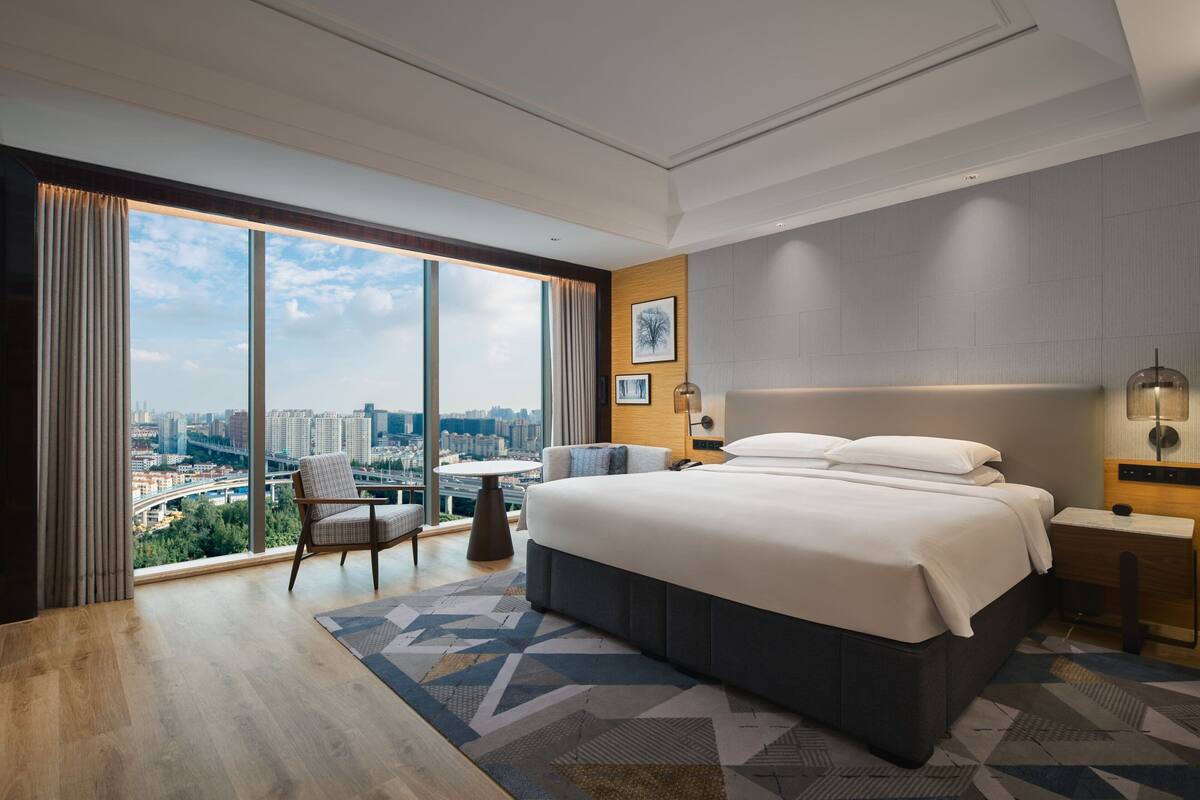 Una habitación de hotel moderna con una cama grande, un escritorio, una silla y vistas panorámicas del paisaje urbano a través de ventanales de piso a techo.