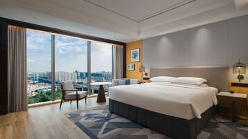 A modern hotel room with a large bed, a desk, a chair, and a view of the cityscape through floor-to-ceiling windows.