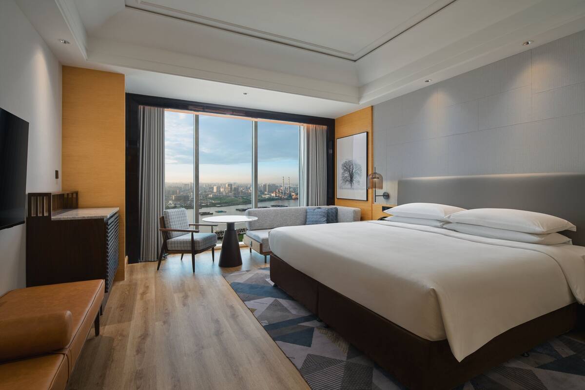 Una habitación de hotel moderna con una cama grande, una zona de estar con una mesa, y vistas a la ciudad a través de amplias ventanas.