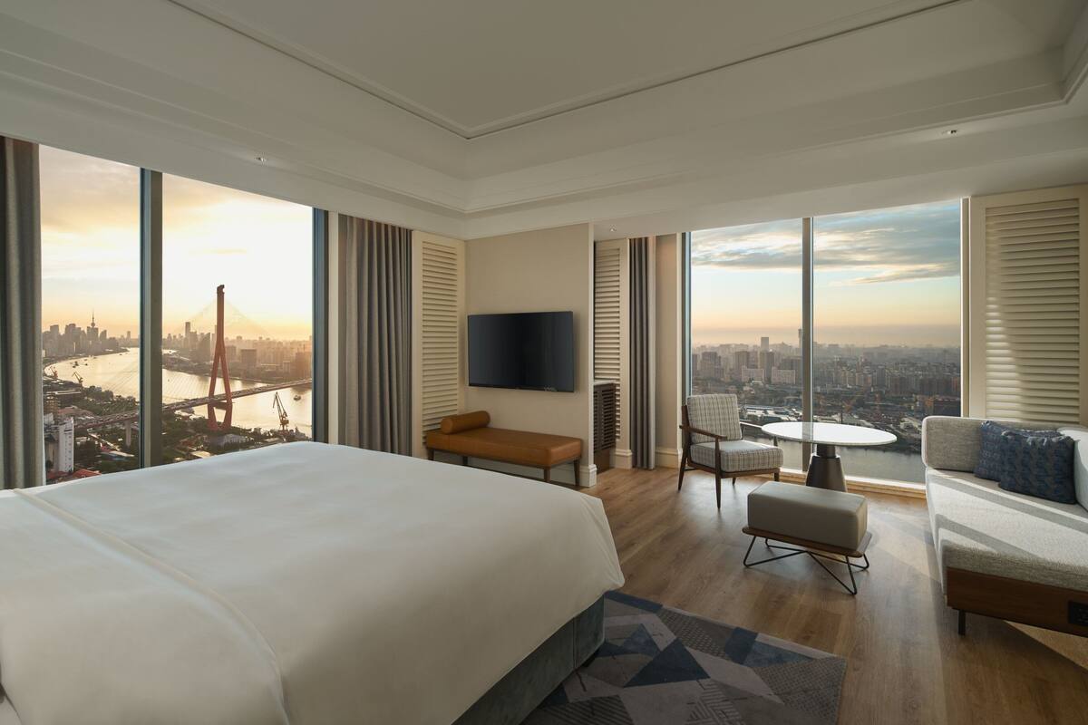 Una habitación de hotel moderna con una cama grande, una zona de estar con un banco y sillas, un televisor de pantalla plana y vistas a la ciudad a través de grandes ventanales.
